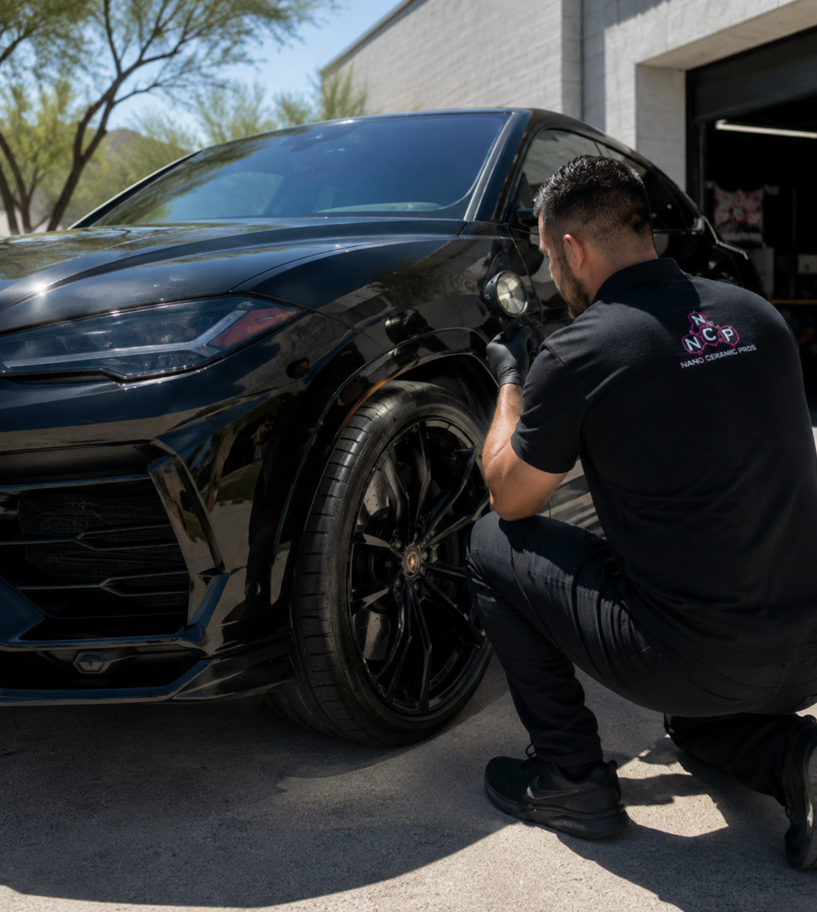 Technician inspecting a coated black luxury SUV under bright outdoor Arizona sunlight with a handheld paint inspection light
